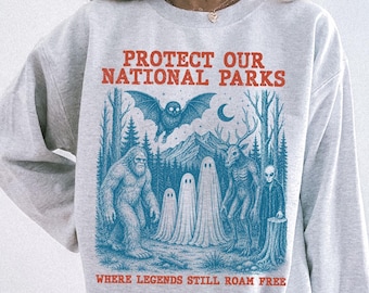 Könnte beinhalten: Ein hellgraues Sweatshirt mit Rundhalsausschnitt und einem blauen Grafikdruck einer Waldszene mit Fledermaus, drei Geistern, einem Sasquatch, einem hirschartigen Wesen und einem Alien. Der Text lautet PROTECT OUR NATIONAL PARKS und WHERE LEGENDS STILL ROAM FREE.