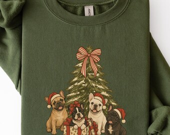 Könnte beinhalten: Ein olivgrüner Rundhals-Pullover mit einem Grafikdruck eines geschmückten Weihnachtsbaums mit einer rot-weiß gestreiften Schleife an der Spitze, umgeben von vier Französischen Bulldoggen mit roten Weihnachtsmützen, eine davon in einer Geschenkbox.