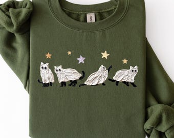 May include: An olive green crewneck sweatshirt featuring an embroidered design of four white cats with black paws and tails, each wearing a white ghost costume. The cats are positioned under five small embroidered stars in gold and light purple.