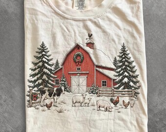 Könnte beinhalten: Ein cremefarbenes T-Shirt mit einem Grafikdruck einer roten Scheune mit Kranz, verschneiten Tannen, einer Kuh, Schafen, einem Schwein und Hühnern. Das Shirt hat ein Comfort Colors Markenetikett am Innenhals.