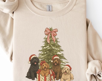 Könnte beinhalten: Ein beigefarbenes Sweatshirt mit Rundhalsausschnitt und einem Aufdruck eines grünen Weihnachtsbaums, der mit einer rot-weiß gestreiften Schleife verziert ist. Vier Hunde mit roten Weihnachtsmützen sitzen davor, einer davon in einer Geschenkbox.