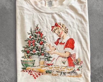 Könnte beinhalten: Ein cremefarbenes T-Shirt mit einem Grafikdruck einer Person in einem roten Kleid und einer weißen Schürze beim Plätzchenbacken. Die Szene zeigt einen geschmückten Weihnachtsbaum, ein Nudelholz, eine Rührschüssel und das Comfort Colors Etikett.