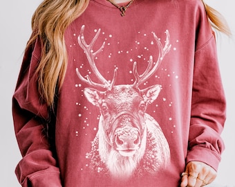 Könnte beinhalten: Ein dunkelrotes Sweatshirt mit Rundhalsausschnitt und einem weißen Grafikdruck eines Rentierkopfes, der von fallenden Schneeflocken umgeben ist. Das langärmelige Kleidungsstück hat eine lockere Passform und eine detaillierte Illustration.