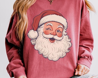 Könnte beinhalten: Ein langärmeliges Sweatshirt mit Rundhalsausschnitt in einem gedämpften Ziegelrot, das einen großen Grafikdruck eines zwinkernden Weihnachtsmanns mit weißem Bart und roter Mütze zeigt. Das Sweatshirt hat eine lockere Passform mit Rippbündchen.