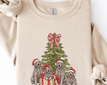 Könnte beinhalten: Ein beigefarbenes Sweatshirt mit Rundhalsausschnitt und einem Aufdruck eines grünen Weihnachtsbaums, der mit einer rot-weiß gestreiften Schleife verziert ist. Vier graue Labrador-Hunde mit roten Weihnachtsmützen sitzen um den Baum herum.