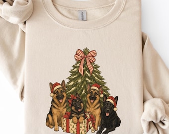 Könnte beinhalten: Ein beigefarbenes Sweatshirt mit Rundhalsausschnitt und einer Grafik von vier Deutschen Schäferhunden mit roten Weihnachtsmützen. Ein Hund sitzt in einer Geschenkbox vor einem Weihnachtsbaum, der mit einer rot-weiß gestreiften Schleife verziert ist.