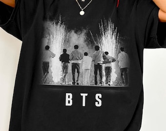 Könnte beinhalten: Ein schwarzes Kurzarm-T-Shirt mit einem Grafikdruck von sieben Figuren, die auf einer Bühne stehen, mit Feuerwerk im Hintergrund. Der Text BTS ist in weißen Großbuchstaben unter dem Bild der Figuren auf der Vorderseite des Shirts gedruckt.