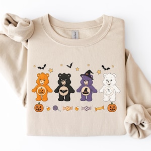 以下が含まれることがあります： ハロウィンの衣装を着た4匹の漫画のクマのグラフィックプリントが施されたベージュのクルーネックトレーナー。クマはオレンジ、黒、紫、白で、カボチャ、コウモリ、魔女の帽子、幽霊が描かれています。クマの周りには小さな星やキャンディが散りばめられています。