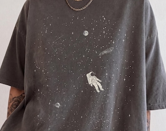 Könnte beinhalten: Ein anthrazitfarbenes Kurzarm-T-Shirt mit einem weißen Grafikdruck eines Astronauten, der im Weltraum zwischen Sternen, einem Mond, einem Planeten mit Ringen und Sternbildern schwebt. Das Shirt hat einen Rundhalsausschnitt und eine lockere Passform.