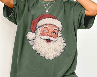 Könnte beinhalten: Ein olivgrünes Kurzarm-T-Shirt mit einem Grafikdruck eines zwinkernden Weihnachtsmanns mit weißem Bart und roter Mütze. Das T-Shirt hat einen Rundhalsausschnitt und eine lockere Passform mit einer detaillierten Illustration.