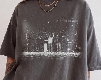 May include: A charcoal gray short-sleeve t-shirt featuring a white graphic print of seven figures standing in a line with arms raised. The text "forever, we are young" is printed in white above the figures, surrounded by small white specks.