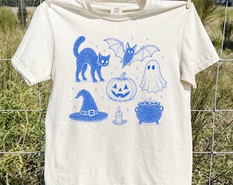 Könnte beinhalten: Ein cremefarbenes Kurzarm-T-Shirt mit einem blauen Grafikdruck, der eine Katze, eine Fledermaus, ein Gespenst, einen Kürbis, einen Hexenhut, eine Kerze und einen Kessel zeigt. Das Design enthält kleine Sternakzente zwischen den Halloween-Motiven.