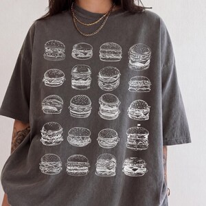 Op de afbeelding: Een antracietgrijs T-shirt met korte mouwen en een witte grafische print van twintig verschillende hamburgerillustraties in een rasterpatroon. Het shirt heeft een ronde hals en een relaxte pasvorm met diverse stijlen hamburgers.