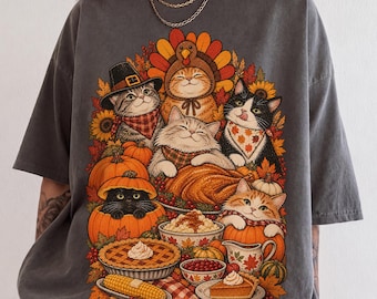 Könnte beinhalten: Ein anthrazitfarbenes Kurzarm-T-Shirt mit einem Grafikdruck von mehreren Katzen, die für Thanksgiving gekleidet sind. Die Katzen sind umgeben von Kürbissen, einem gebratenen Truthahn, einem Kuchen, Mais und Herbstblättern auf einer karierten Tischdecke.