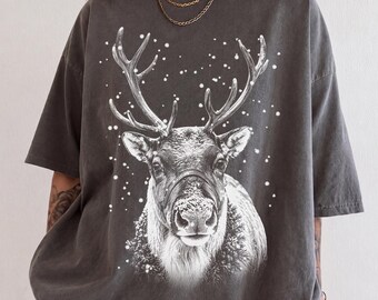 Könnte beinhalten: Ein anthrazitfarbenes Kurzarm-T-Shirt mit einem großen weißen Grafikdruck eines Rentierkopfes mit Geweih, umgeben von fallendem Schnee. Das Rentier ist mit detaillierter Fellstruktur und einer verschneiten Nase auf der Brust abgebildet.