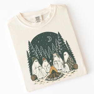 May include: A cream-colored t-shirt featuring a graphic of four ghosts sitting around a campfire in a forest at night. The ghosts wear hats and hold mugs or a marshmallow on a stick. The design includes pine trees, a crescent moon, and stars.