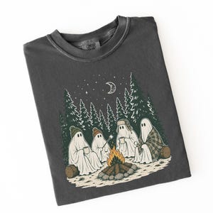 May include: A charcoal gray crew neck t-shirt featuring a graphic illustration of four ghosts sitting around a campfire in a forest at night. The ghosts wear hats and blankets, holding mugs and a marshmallow on a stick under a crescent moon.