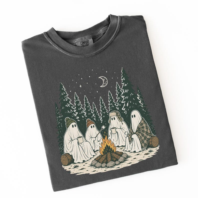 May include: A charcoal gray crew neck t-shirt featuring a graphic illustration of four ghosts sitting around a campfire in a forest at night. The ghosts wear hats and blankets, holding mugs and a marshmallow on a stick under a crescent moon.