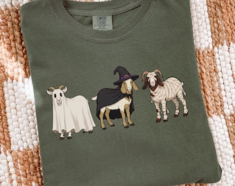 May include: An olive green crew neck t-shirt featuring a graphic of three goats in costumes. One goat is dressed as a ghost, one as a witch with a black cape and hat, and one as a mummy wrapped in white bandages. The shirt has a Comfort Colors label.