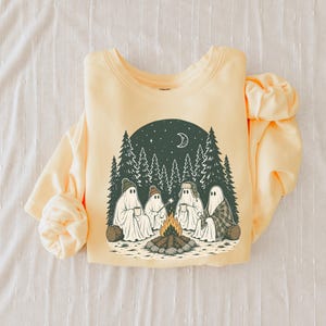 May include: A pale yellow crewneck sweatshirt featuring a graphic print of four ghosts sitting around a campfire in a forest. The scene includes pine trees, a crescent moon, stars, and the ghosts wearing hats and holding mugs and a roasting stick.