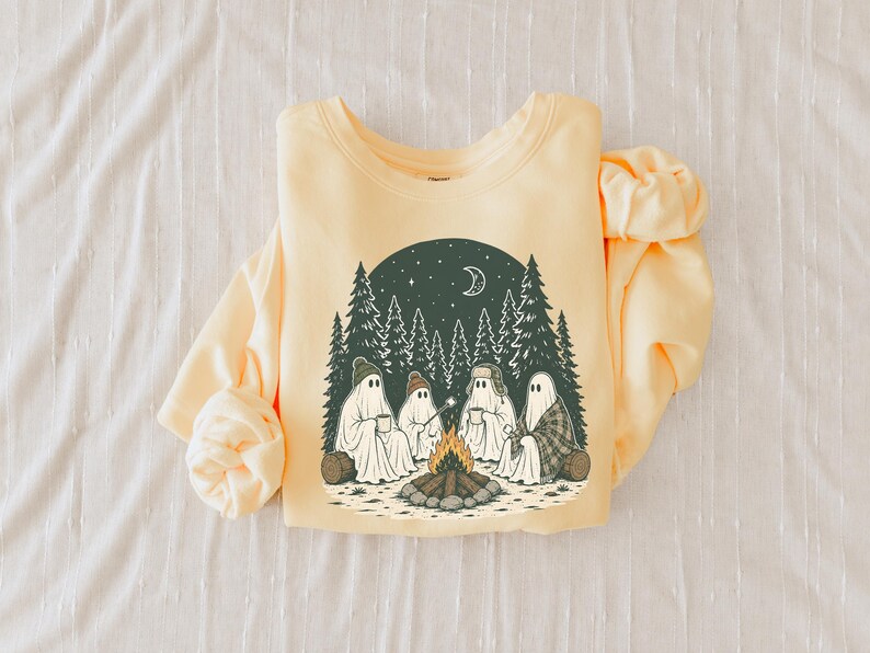 May include: A pale yellow crewneck sweatshirt featuring a graphic print of four ghosts sitting around a campfire in a forest. The scene includes pine trees, a crescent moon, stars, and the ghosts wearing hats and holding mugs and a roasting stick.