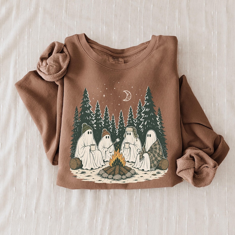 May include: A brown long-sleeve sweatshirt featuring a graphic print of four ghosts sitting around a campfire in a forest. The ghosts wear hats and hold mugs, with a crescent moon and stars above the trees. The design includes pine trees and logs.