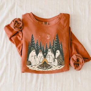 May include: A rust-colored crewneck sweatshirt featuring a graphic print of four ghosts sitting around a campfire in a pine forest. The illustration includes a crescent moon, stars, and the ghosts wearing hats and holding mugs under the night sky.