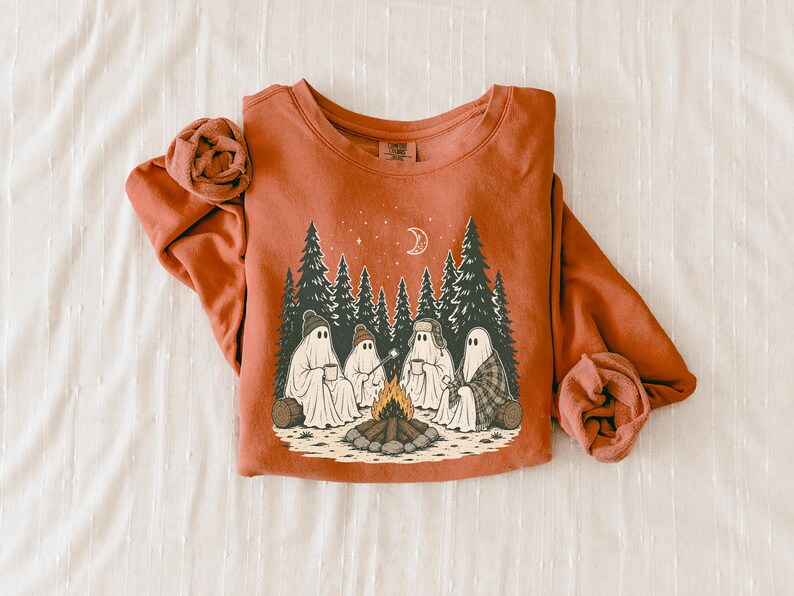 May include: A rust-colored crewneck sweatshirt featuring a graphic print of four ghosts sitting around a campfire in a pine forest. The illustration includes a crescent moon, stars, and the ghosts wearing hats and holding mugs under the night sky.