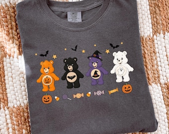 May include: A charcoal gray crew neck t-shirt featuring a graphic print of four cartoon bears with Halloween themes, including a pumpkin, bat, witch hat, and ghost. The design includes small stars, bats, and candy illustrations. The label reads Comfort Colors.