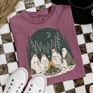 May include: A maroon t-shirt featuring a graphic of four ghosts sitting around a campfire in a forest at night. The ghosts wear hats and hold mugs, with a crescent moon and stars in the dark green sky above the trees. The design includes a log fire.