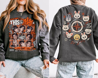 Könnte beinhalten: Ein anthrazitfarbenes Langarm-Sweatshirt mit einer Vorderseite, die Fotos der Stray Kids-Mitglieder in einem Raster, orangefarbene Akzente und den Text "THIS & THAT" zeigt. Die Rückseite zeigt ein kreisförmiges Kompassdesign mit Tierfiguren.