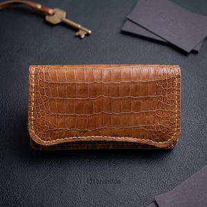 Peut inclure: Un étui à clés rectangulaire en cuir marron avec un motif texturé en peau de crocodile et des coutures claires visibles sur les bords. L'étui est fermé et placé horizontalement, accompagné d'une clé en métal et de cartes noires.