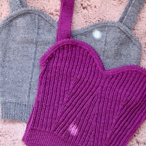 Op de afbeelding: Twee gebreide tanktops, één felpaars en één grijs. Beide hebben een ronde hals en brede bandjes. De paarse top heeft een geribbelde textuur. De tops worden gepresenteerd op een roze, fuzzy oppervlak.