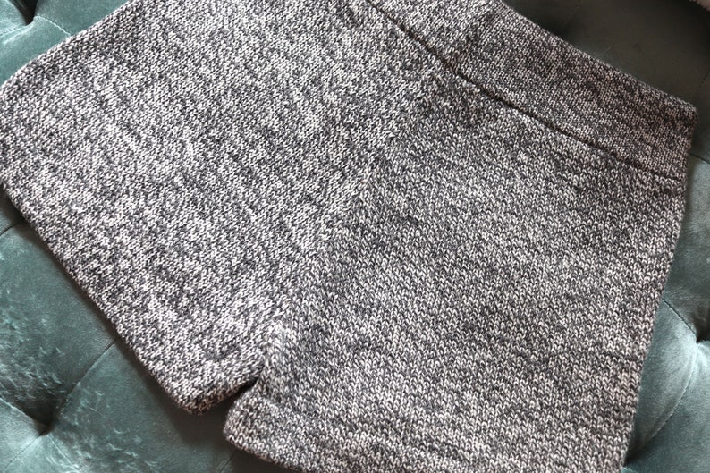 May include: Close-up of gray knit shorts. The shorts have a textured appearance with a mix of light and dark gray threads. The garment is folded, showcasing the waistband and the fabric's detailed weave. The shorts appear to be made of a soft, comfortable material.