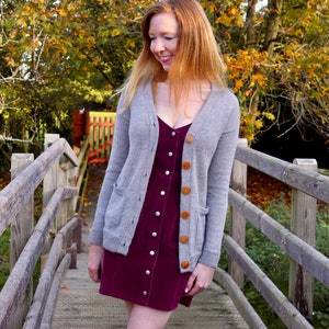 Può includere: Una donna indossa un abito in velluto a coste bordeaux con un cardigan grigio e bottoni marroni. È in piedi su un ponte di legno con uno sfondo di fogliame autunnale.