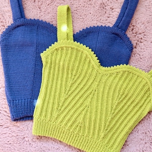 May include: Three knitted crop tops in shades of blue and chartreuse. The tops have a scoop neckline and wide straps. The chartreuse top has a ribbed chevron design. The tops are displayed on a pink, textured surface.