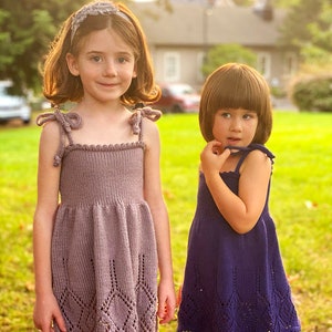 Puede incluir: Dos niñas pequeñas con vestidos de punto a juego con un patrón de encaje. La niña de la izquierda lleva un vestido morado claro y una diadema. La niña de la derecha lleva un vestido morado oscuro.