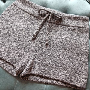 May include: A pair of gray and white knit shorts with a drawstring waist. The shorts are made of a textured fabric and have a casual, comfortable style. The shorts are shown on a teal, tufted surface.