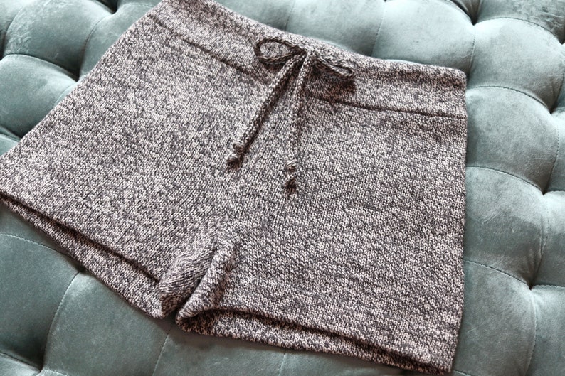 May include: A pair of gray and white knit shorts with a drawstring waist. The shorts are made of a textured fabric and have a casual, comfortable style. The shorts are shown on a teal, tufted surface.
