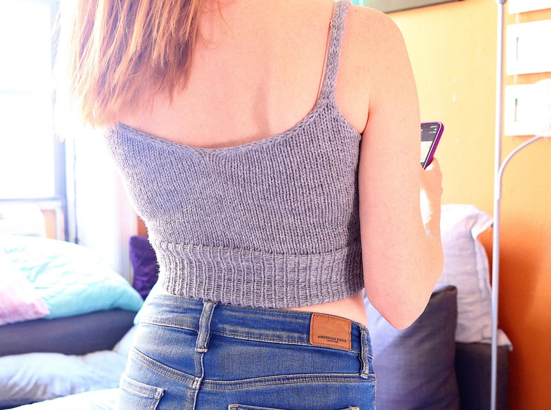 May include: A gray knitted crop top with thin straps. The top is worn with blue jeans and the label "American Eagle" is visible on the back of the jeans.