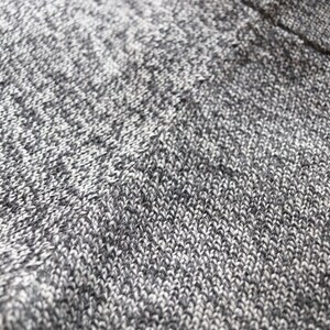 May include: Close-up of a knitted fabric with a speckled pattern. The textile is primarily gray and white, with a subtle texture created by the knit stitches. The fabric appears soft and possibly warm, suitable for clothing or home decor.