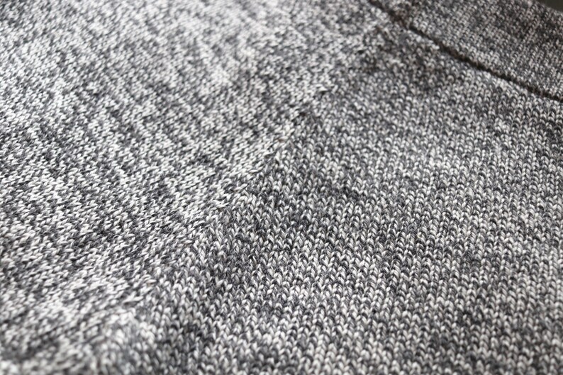 May include: Close-up of a knitted fabric with a speckled pattern. The textile is primarily gray and white, with a subtle texture created by the knit stitches. The fabric appears soft and possibly warm, suitable for clothing or home decor.