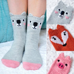 Könnte beinhalten: Ein Paar graue Stricksocken mit rosa Zehen und Fersen. Jede Socke hat ein weiß-schwarzes gesticktes Tiergesicht. Die Tiere sind ein Bär, ein Fuchs und ein Schwein.