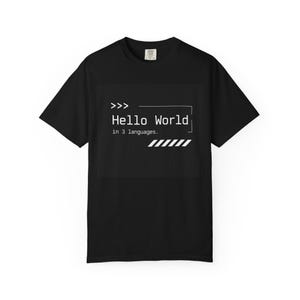 以下が含まれることがあります： 胸元に白いグラフィックプリントが施された黒の半袖Tシャツ。デザインには、3つの矢印、「Hello World in 3 languages.」というテキスト、長方形の枠線、右下の角に一連の斜めのストライプが含まれています。