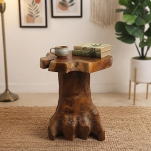 May include: A natural wood side table crafted from a tree stump with a smooth, polished top surface. A ceramic mug and two stacked hardcover books with light-colored covers are placed on the table. The wood features a rich brown grain.