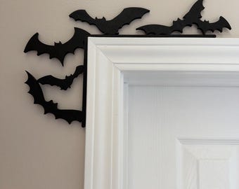 May include: A black, L-shaped decorative wall accent featuring a series of bat silhouettes. The piece is designed to fit snugly around the top corner of a white door frame, with two bats extending vertically down the side and three bats horizontally.