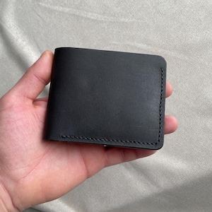 May include: A black leather bifold wallet featuring a smooth, matte finish and visible dark stitching along the edges. The wallet has a rectangular shape with rounded corners and a clean, simple design that highlights the texture of the material.