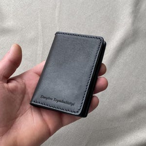 May include: A black leather card holder featuring visible stitching along the edges and the name Dmytro Tsymbalistyi embossed in the bottom left corner. The rectangular accessory has a smooth matte finish and a structured, folded design.