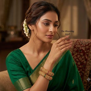May include: Two gold bangles with intricate textured patterns and small green and red stone accents worn on a wrist. The jewelry is paired with a deep green garment featuring a gold border. The text The Blooming Jewel is visible in the image.