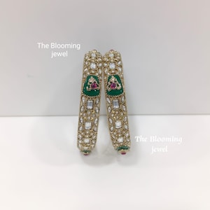Op de afbeelding: Twee goudkleurige armbanden met ingewikkeld metaalwerk, heldere rechthoekige en ronde stenen, en een centraal groen geëmailleerd gedeelte met een kleine roze steen. De tekst The Blooming jewel is zichtbaar in de afbeelding.